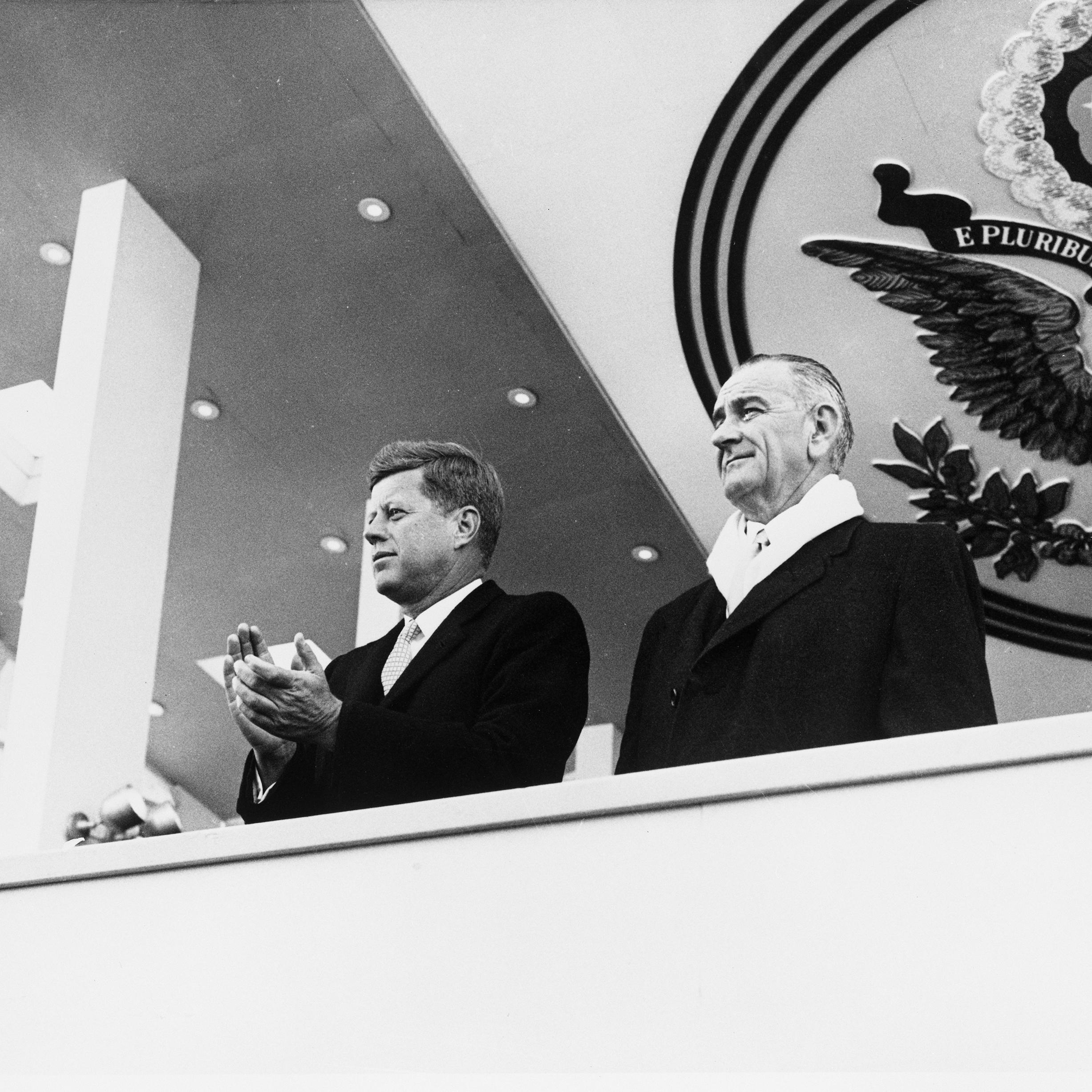 President Kennedy and Vice President Johnson watch their inauguration parade from a balcony, with the United States seal behind and above them.